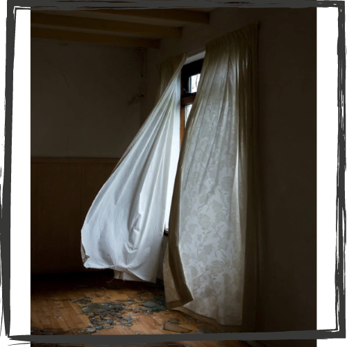Photo of a window with long, white curtains billowing in the draft because the glass is broken and laying on the bare, wood floor