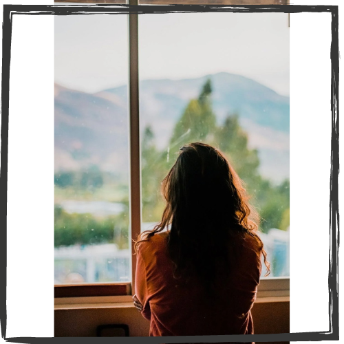 Photo of a woman with long hair sitting looking out a large window. Her back is to the camera.