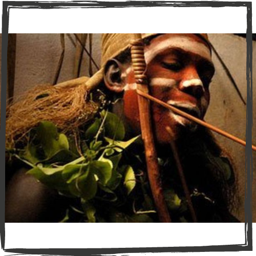 An Gabonese man with his brow, nose, cheeks & lips painted white and a headcovering made of a brown, woven material, uses a rod & stick to administer iboga