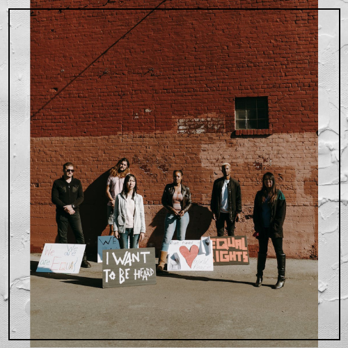 6 white, Asian, Black and bi-racial people stand near a brick wall with signs about racism at their feet
