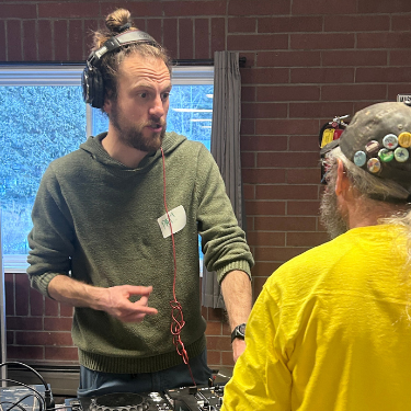 A young, white man with hair in a bun & wearing headphones chats with a bearded man in a yellow shirt