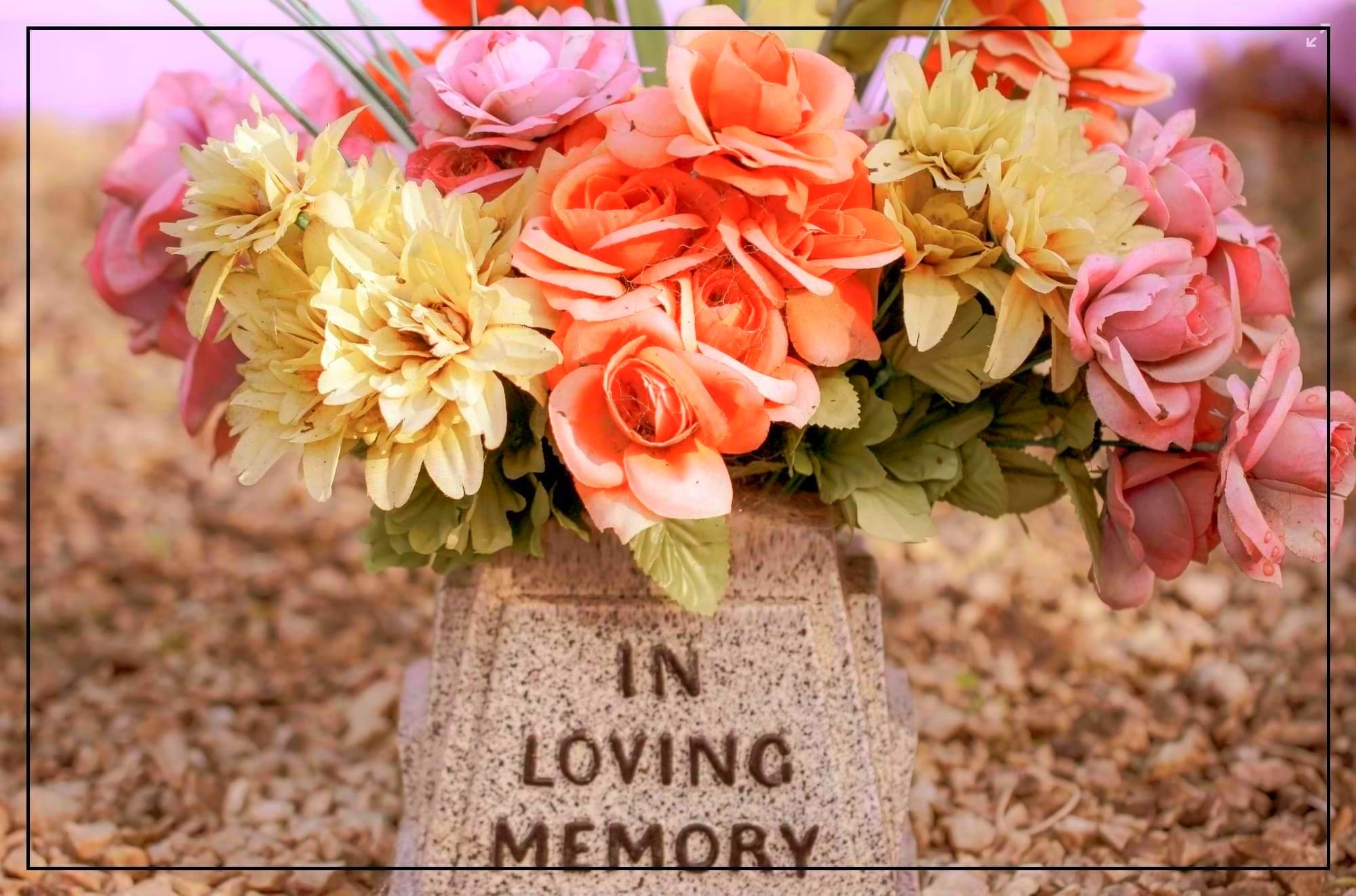 Photo of a burial marker with the words, "In Loving Memory" and covered with multi-colored flowers