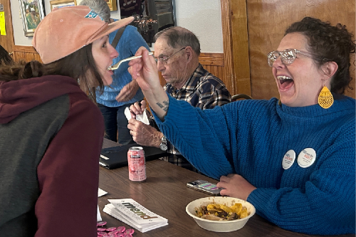 One white woman in a pink cap gets a spoonful of food from a seated woman laughing